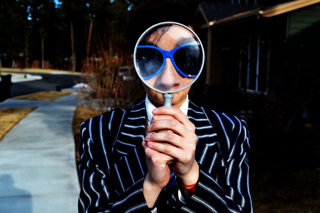 Man in a suit and blue-rimmed sunglasses looks through a magnifying glass