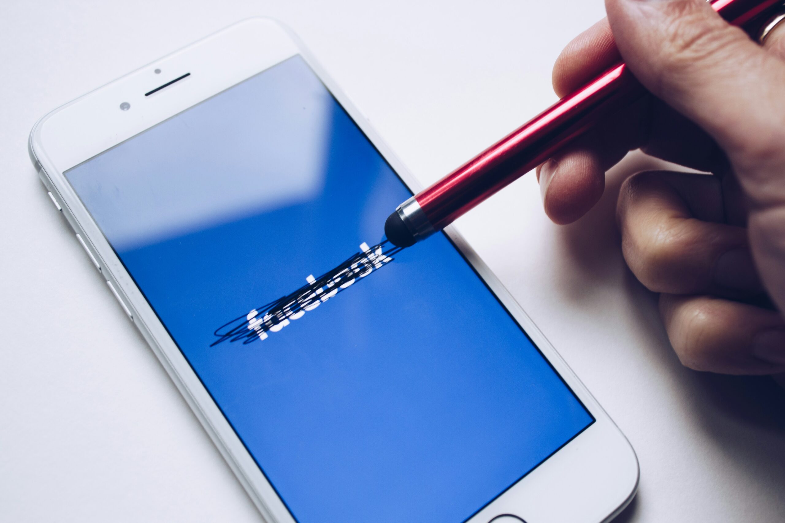 A stylus held by a hand hovering over a smartphone with the Facebook logo scribbled out on it